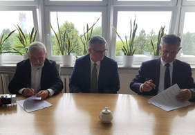 Trzech mężczyzn siedzi za stołem nad dokumentami, za nimi ściana okien