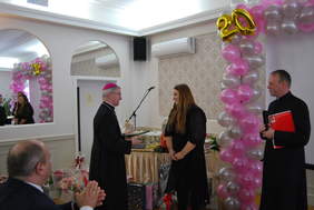 Między dwoma duchownymi stoi pani kierownik, biskup jest skierowany w jej stronę, mówi do niej