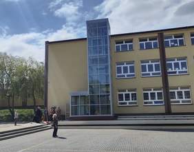 Pokaż zdjęcie: Duży jasny budynek szkolny z wielkimi oknami, z białą ramą. Do niego przymocowana szklana konstrukcja z winą w środku. Przed budynkiem plac z kostki na którym stoi kobieta.