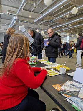 Pokaż zdjęcie: Zdjęcie zrobione zza stoiska wystawienniczego PFRON, widać stolik na którym leżą ulotki oraz plecy kobiety w czerwonym golfie.