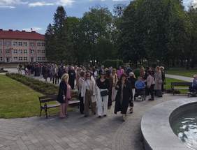 Pokaż zdjęcie: Grupa ludzi spaceruje alejką w parku obok fontanny. W tle budynek z czerwonym dachem, dużo zieleni i drzew. Na niebie widoczne rozproszone chmury, słoneczna pogoda.