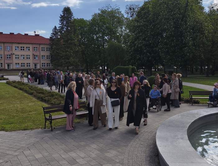 Pokaż zdjęcie: Grupa ludzi spaceruje alejką w parku obok fontanny. W tle budynek z czerwonym dachem, dużo zieleni i drzew. Na niebie widoczne rozproszone chmury, słoneczna pogoda.