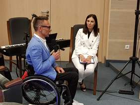 kobieta w białym garniturze, siedzi z prawej strony, prowadzi wywiad z mężczyzna w niebieskiej marynarce, który siedzi na wózku inwalidzkim