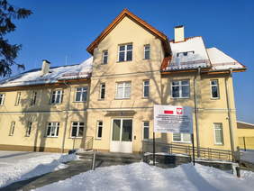 Pokaż zdjęcie: Dwupiętrowy, jasny budynek z czerwonym dachem, stojący wśród śniegu. Do głównego wejścia prowadzą schody i podjazd z poręczą dla osób z niepełnosprawnościami; przed budynkiem ustawiona jest tablica informacyjna.