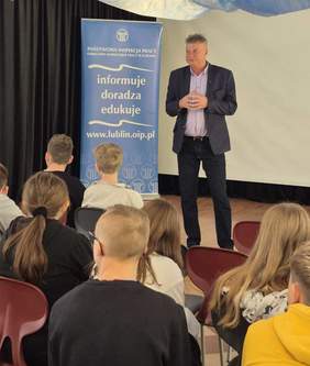 Pokaż zdjęcie: Do dzieci zgromadzonych na sali gimnastycznej przemawia mężczyzna w marynarce, w ręku trzyma mikrofon.