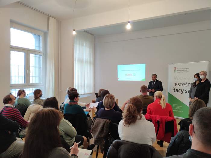 Pokaż zdjęcie: Fragment sali, od tyłu siedzący uczestnicy. Z przodu Sali przodem do słuchaczy od lewej: Jan Wroński Dyrektor Oddział Śląskiego PFRON. Z prawej na tle LOGO PFRON - Teresa Saczuk-Grabowska i Ewa Samełko - pracownicy PFRON.