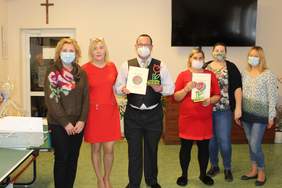 Pokaż zdjęcie: Sześć osób- pięć kobiet i jeden mężczyzna. Mężczyzna i kobieta stojący na środku trzymają dyplomy i statuetki przedstawiające czerwono-zielonego kwiatka. Z tyłu na ścianie widzimy wiszący czarny telewizor. Szarowola Zdjęcie 8140