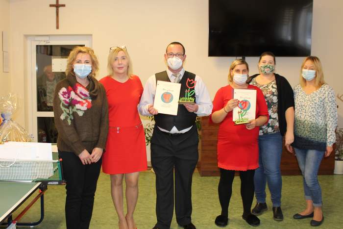 Pokaż zdjęcie: Sześć osób- pięć kobiet i jeden mężczyzna. Mężczyzna i kobieta stojący na środku trzymają dyplomy i statuetki przedstawiające czerwono-zielonego kwiatka. Z tyłu na ścianie widzimy wiszący czarny telewizor. Szarowola Zdjęcie 8140