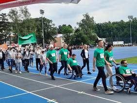 Pokaż zdjęcie: Po boisku olimpijskim idzie tłum ludzi. Niektórzy jadą na ręcznych wózkach inwalidzkich. Są ubrani w sportowe stroje i niosą transparenty. 