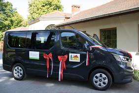 Pokaż zdjęcie: Czarny mini bus oznakowany logo PFRON i DPS w Radawiu. Klamki przewiązane kokardami z czerwono-białą szarfą. W tle fragment budynku