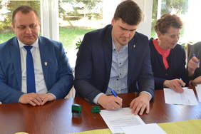 Pokaż zdjęcie: Na zdjęciu znajduje się dwóch mężczyzn i kobieta. Młody mężczyzna podpisuje umowę