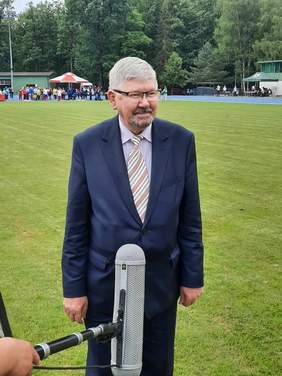 Pokaż zdjęcie: Przed mikrofonem telewizyjnym stoi mężczyzna. Jest ubrany w garnitur i się uśmiecha. W tle widać zieloną murawę i zawodników sportowych. 