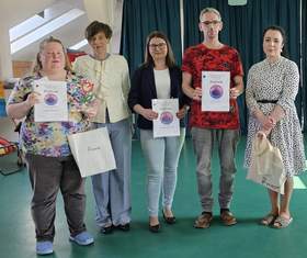 Na zdjęciu od lewej: kobieta w kwiecistej bluzce i niebieskich klapkach, trzymająca dyplom, białą torbę PFRON oraz statuetkę tulipana. Obok niej kobieta z krótkimi ciemnymi włosami w jasnej marynarce, a następnie kobieta w okularach i czarnej marynarce, trzymająca dyplom. Kolejną osobą jest mężczyzna w czerwonej koszulce z dyplomem w dłoni. Grupę zamyka kobieta w białej sukience w grochy, trzymająca białą torbę z logo PFRON.