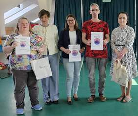 Pokaż zdjęcie: Na zdjęciu od lewej: kobieta w kwiecistej bluzce i niebieskich klapkach, trzymająca dyplom, białą torbę PFRON oraz statuetkę tulipana. Obok niej kobieta z krótkimi ciemnymi włosami w jasnej marynarce, a następnie kobieta w okularach i czarnej marynarce, trzymająca dyplom. Kolejną osobą jest mężczyzna w czerwonej koszulce z dyplomem w dłoni. Grupę zamyka kobieta w białej sukience w grochy, trzymająca białą torbę z logo PFRON.