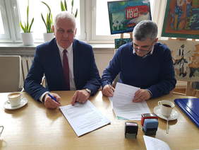 Dwaj mężczyźni siedzący przy jasnym stole na tle okien, podpisują dokumenty, na stole są też pieczątki, stoją kawy