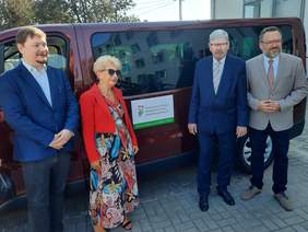 Pokaż zdjęcie: Przed samochodem stoi kobieta i trzech mężczyzn. Na busie naklejona jest naklejka z informacją, że auto zakupiono ze środków PFRON. 