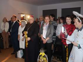 Pokaż zdjęcie: Zdjęcie gości, którzy stoją podczas uroczystości, na pierwszym planie kobieta blondynka z koszem cukierków oraz biskup. Z boku stoi mężczyzna anioł.