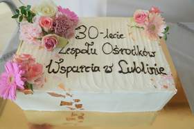 Prostokątny biały tort jubileuszowy ozdobiony różowymi kwiatami. Na torcie napis z czekolady: „30‑lecie Zespołu Ośrodków Wsparcia w Lublinie”. 