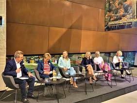 Pokaż zdjęcie: Mężczyzna i pięć kobiet siedzą w fotelach w rzędzie. Mężczyzna trzyma mikrofon i mówi coś w stronę kobiet.