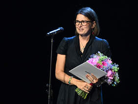 Pokaż zdjęcie: Kobieta w okularach stoi przy mikrofonie, trzyma bukiet kwiatów i dokument z logo „gdynia.pl”. Ubrana w elegancką, ciemną sukienkę, uśmiecha się w stronę publiczności. Tło ciemne, scena dobrze oświetlona.