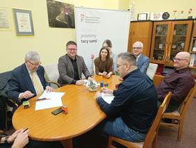 Pokaż zdjęcie: Przy owalnym stole siedzi kobieta i pięciu mężczyzn. Podpisują umowę. Na stole leżą różne dokumenty, pieczątki, telefony. 