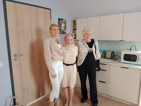 Pokaż zdjęcie: W kuchni stoją trzy kobiety, które pozują do zdjęcia. Są uśmiechnięte. W tle widać sprzęty kuchenne. 