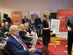 Pokaż zdjęcie: Marlena Maląg, Prezes Zarządu PFRON prezentuje okolicznościową kartkę i znaczek pocztowy wydany przez Pocztę Polską