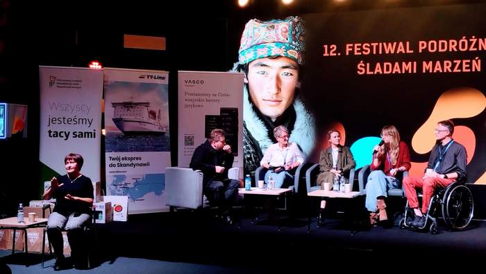 Pokaż zdjęcie: Pięć osób siedzi na tle dużego ekranu z napisem: „12. Festiwal Podróżniczy Śladami Marzeń” i zdjęciem mężczyzny w kożuchu, z wzorzystą czapką na głowie. Z przodu, na tle baneru PFRON oraz dwóch innych banerów siedzi tłumaczka języka migowego.