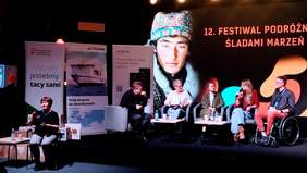 Pokaż zdjęcie: Pięć osób siedzi na tle dużego ekranu z napisem: „12. Festiwal Podróżniczy Śladami Marzeń” i zdjęciem mężczyzny w kożuchu, z wzorzystą czapką na głowie. Z przodu, na tle baneru PFRON oraz dwóch innych banerów siedzi tłumaczka języka migowego.