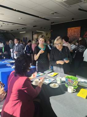 Pokaż zdjęcie: Przy stole siedzi kobieta, na stole leżą ulotki, po które sięgają zainteresowani.