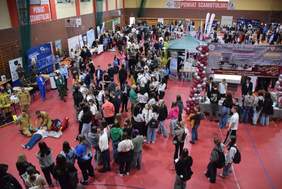 Pokaż zdjęcie: Liczna grupa młodzieży znajduje się w hali sportowej. W hali można dostrzec dużą liczbę wystawców wraz z banerami. Po środku znajduje się namiot jednego z wystawców.