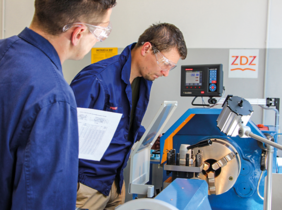 Pokaż zdjęcie: Przy maszynie stoi dwóch mężczyzn, ubranych w niebieskie stroje robocze. Mają okulary. Przyglądają się maszynie