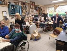 Pokaż zdjęcie: Przy stołach stolarskich i piłach siedzą kilku uczestników warsztatów. Osoba na wózku w maseczce patrzy w obiektyw aparatu, tuż za nią mężczyzna odpowiada na pytanie zadane przez panią dyrektor. Oboje są zwróceni do siebie. W dali instruktor 