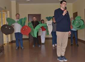 Pokaż zdjęcie: Na pierwszym planie mężczyzna trzyma mikrofon w ręku i śpiewa. Za nim 5 osób trzyma rysunki warzyw