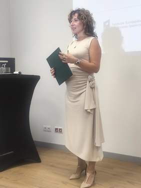 Pokaż zdjęcie: Osoba w jasnej, eleganckiej sukni stoi przy stoliku i przemawia, trzymając zieloną teczkę; w tle ekran projekcyjny i wnętrze sali konferencyjnej.