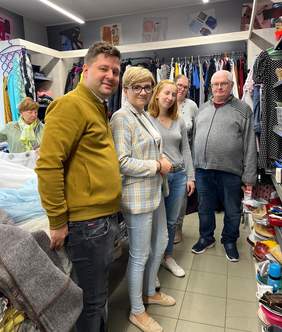 Pokaż zdjęcie: Pięć osób w sklepie charytatywnym, otoczonych półkami i wieszakami pełnymi ubrań.