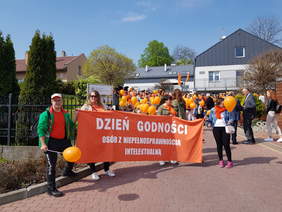 Jasny, słoneczny dzień. Uczestnicy marszu z pomarańczowymi balonami i transparentem zbierają się przed siedzibą PSONI