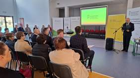 Pokaż zdjęcie: Duża sala. Na krzesłach ułożonych w rzędach siedzi grupa ludzi. Widownia obserwuje prezentację wyświetlaną wyświetlaną na dużym ekranie. W tle prelegent stoi z mikrofonem przed ekranem.