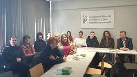 Pokaż zdjęcie: Kilkanaście osób siedzi przy długim białym stole z rozłożonymi ulotkami. Na ścianie za uczestnikami wisi tablica z napisem „Państwowy Fundusz Rehabilitacji Osób Niepełnosprawnych”. Po lewej stronie zdjęcia widać okno z pionowymi żaluzjami.