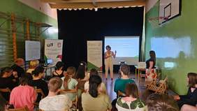 Pokaż zdjęcie: Zdjęcie z tyłu sali w kierunku prelegentki, która wskazuje ręką na ekran z prezentacją.