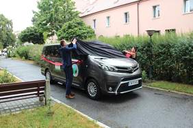 Mikrobus marki Toyota, udekorowany czerwonymi wstążkami, odkrywany z pokrowca przez dwie osoby