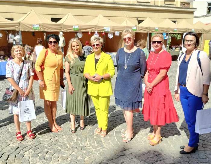 Pokaż zdjęcie: Na rynku starego miasta do pamiątkowego zdjęcia pozuje 7 kobiet ubranych w letnie kolorowe stroje, w tle stoiska wystawców. 