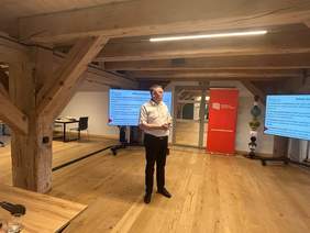 Na środku sali stoi prelegent - przedstawiciel Kujawsko-Pomorskiego Oddziału PFRON. W tle, po bokach, stoją dwa ekrany z wyświetlona prezentacją, a na środku baner Pracodawców Pomorza i Kujaw.