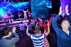 Pokaż zdjęcie: Fotografia przedstawia zarówno widownię jak i scenę. Widownia na pierwszym planie ma uniesione ręce bijące brawo. Na scenie zespół muzyczny, wokalista śpiewający do mikrofonu ma uniesione ręce. Trzech mężczyzna gra na gitarach a w tle widać perkusistę. Cała scena oświetlona jest światłami w niebieskim kolorze. 