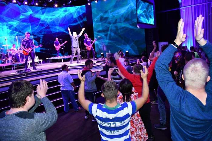 Pokaż zdjęcie: Fotografia przedstawia zarówno widownię jak i scenę. Widownia na pierwszym planie ma uniesione ręce bijące brawo. Na scenie zespół muzyczny, wokalista śpiewający do mikrofonu ma uniesione ręce. Trzech mężczyzna gra na gitarach a w tle widać perkusistę. Cała scena oświetlona jest światłami w niebieskim kolorze. 