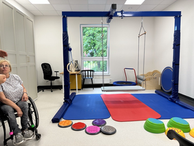 Pokaż zdjęcie: W sali znajduje się osoba na wózku, na środku Sali duży stelaż metalowy pod nim kolorowe maty do ćwiczeń. Po lewej stronie duża szafa.