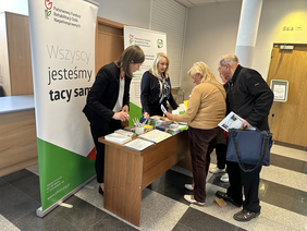 Pokaż zdjęcie: Przy biurku stoją dwie kobiety ubrane w marynarkach. Na biurku materiały promocyjne. Za nimi dwa banery. Jeden z logo PFRON i napisem wszyscy jesteśmy tacy sami. Drugi z logo PFRON i CIDON. Przed nimi stoją ludzi biorący ulotki.