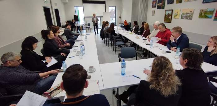 Widok na salę konferencyjną. Na pierwszym planie uczestnicy siedzący przy stole, przed nimi stoi i przemawia p. Łukasz Żmuda - Dyrektor Oddziału Opolskiego PFRON 