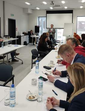 Widok na salę konferencyjną. Na pierwszym planie uczestnicy siedzący przy stole, przed nimi stoi i przemawia p. Łukasz Żmuda - Dyrektor Oddziału Opolskiego PFRON 