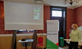 Pokaż zdjęcie: Kobieta w zielonej bluzie z mikrofonem w dłoni przemawia na ekranie wyświetla się prezentacja. 